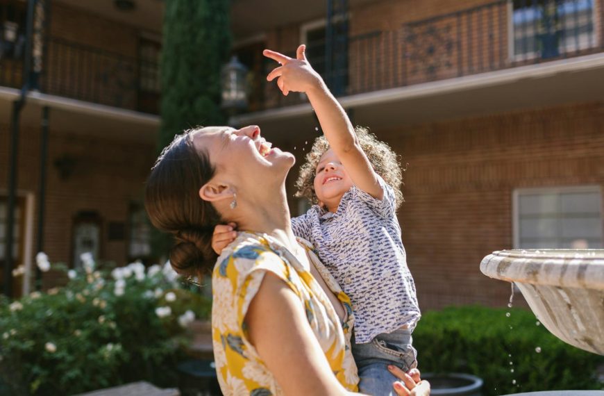 A mother and child share a joyful moment by a water fountain in a sunny garden setting.