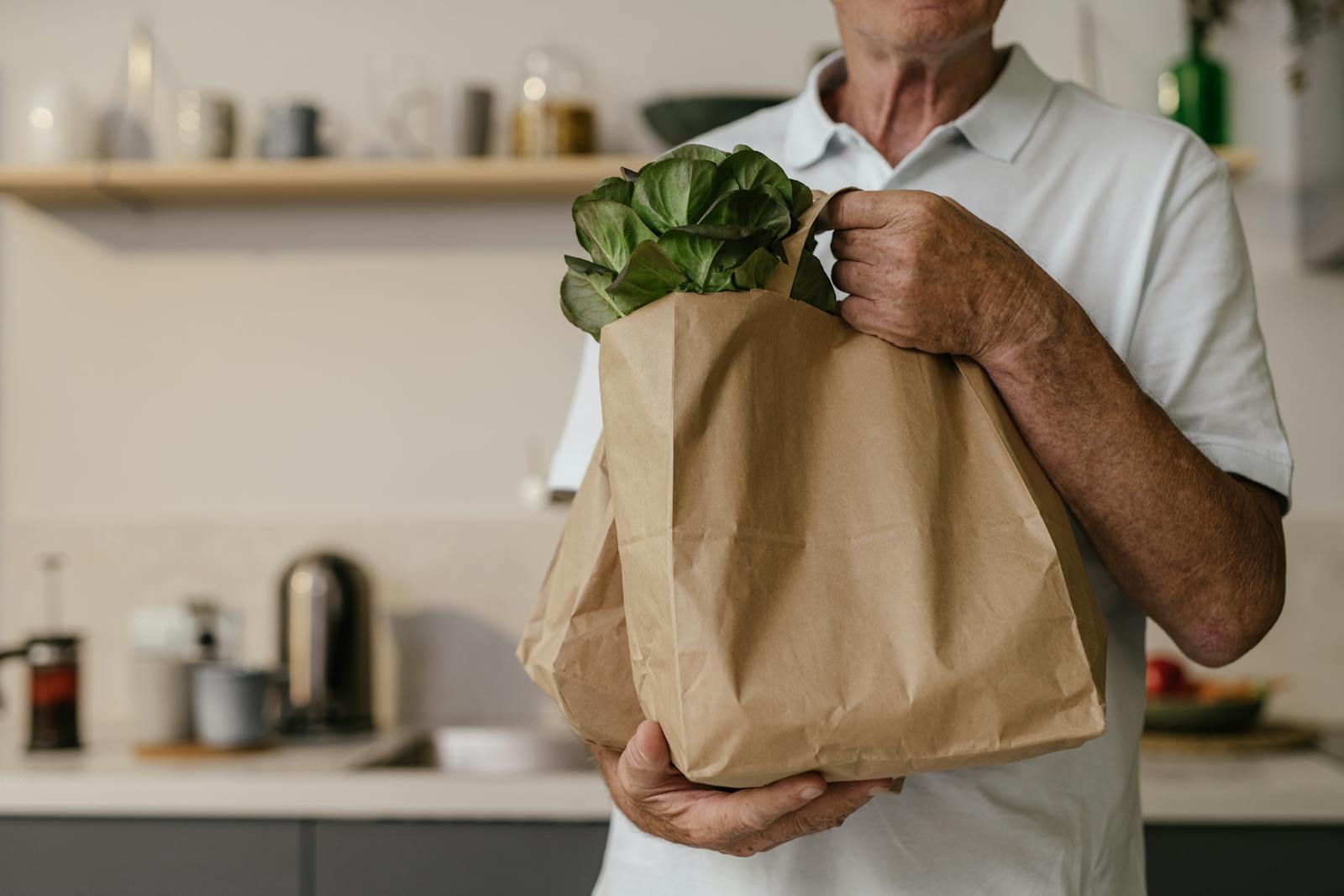 An older man holds a paper bag filled with fresh vegetables in a kitchen, conveying a healthy lifestyle.