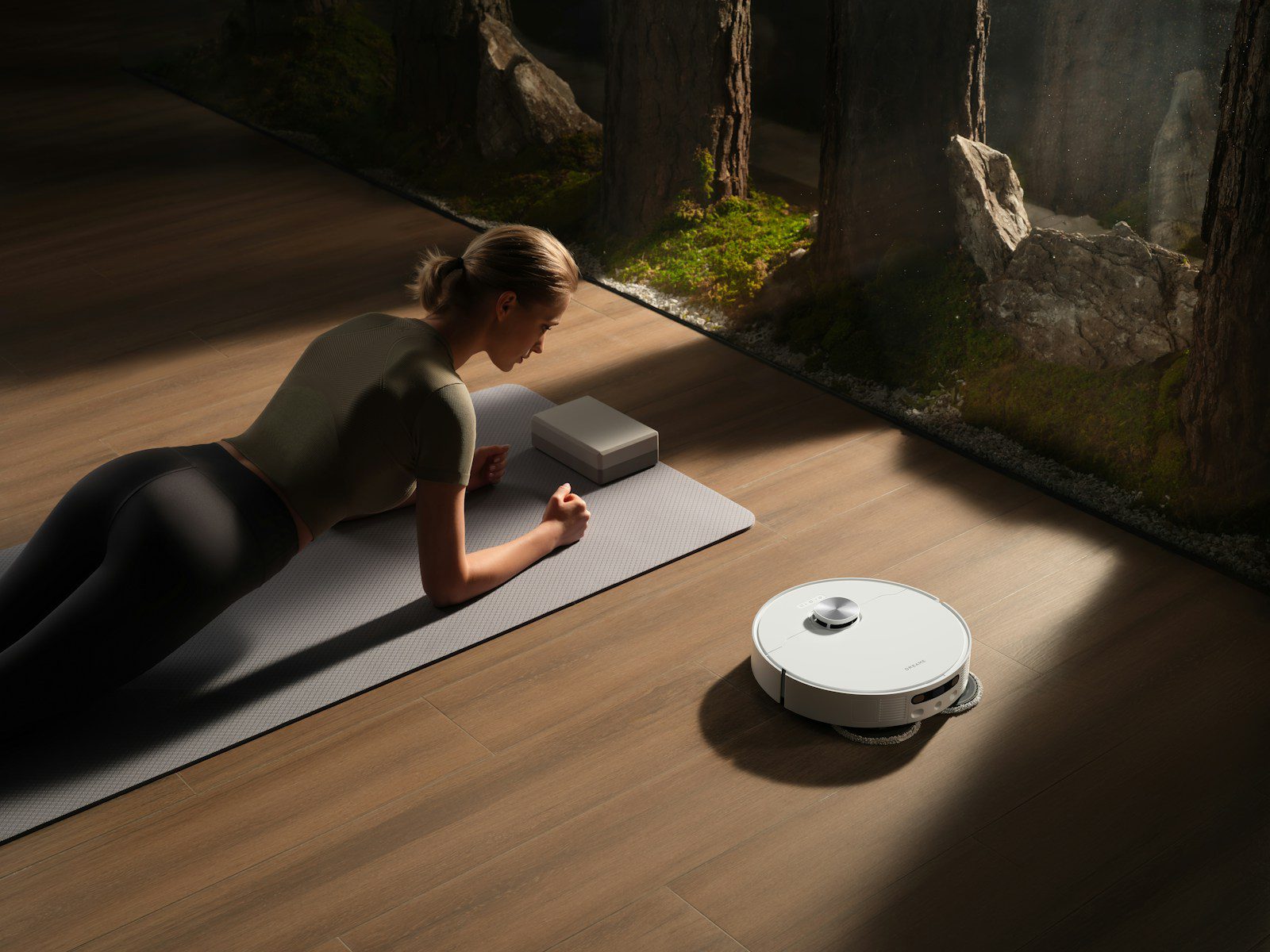 Woman planks on mat near robot vacuum cleaner.