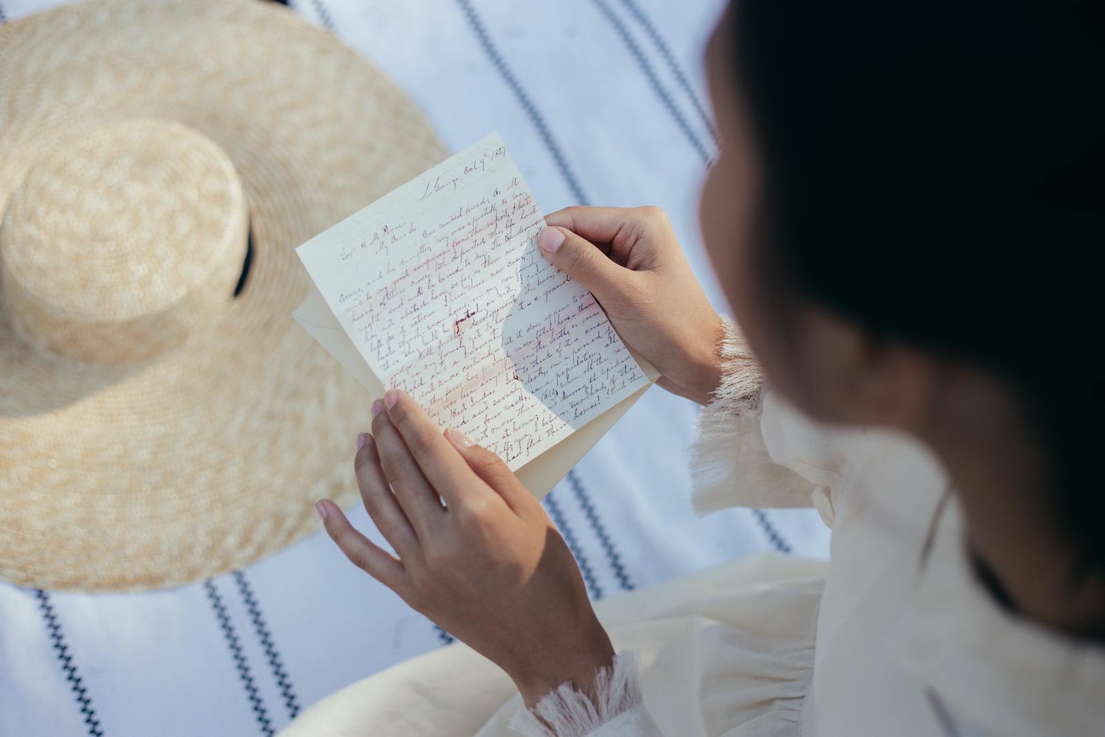 A young woman reads a handwritten love letter while seated outdoors beside a straw hat.
