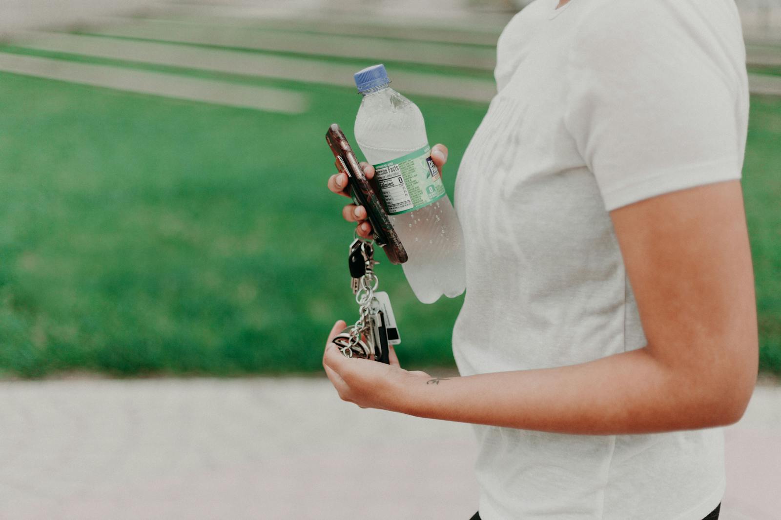 A person holding a water bottle, phone, and keys while walking outdoors.