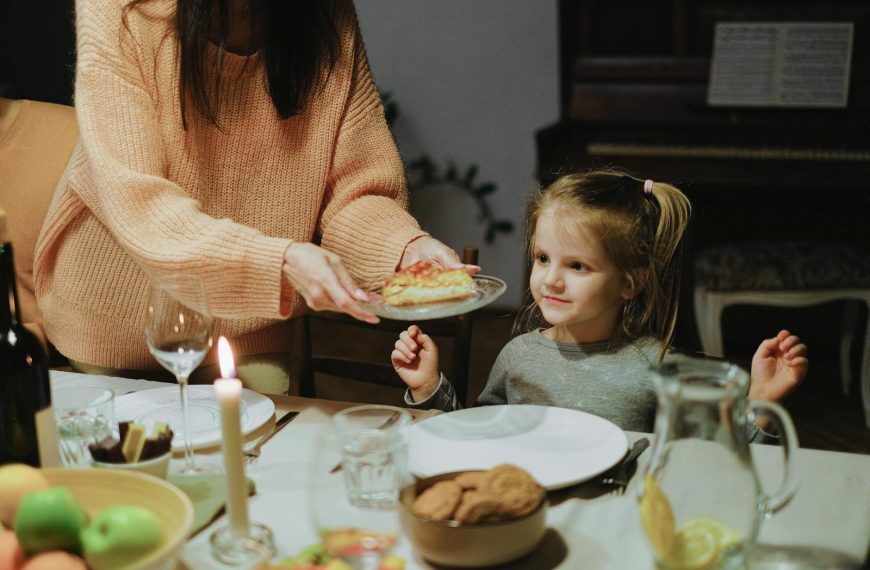 A mother serves dinner to her young daughter at a warmly lit family table, creating a cozy atmosphere.