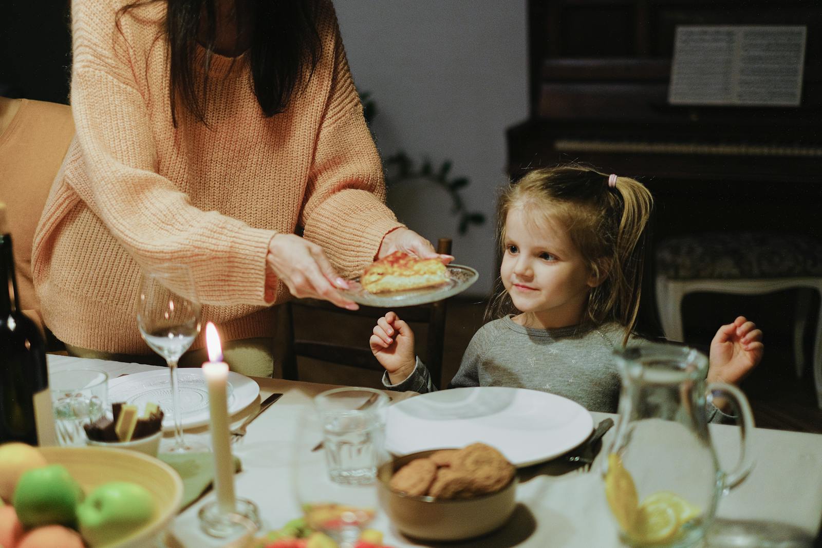 A mother serves dinner to her young daughter at a warmly lit family table, creating a cozy atmosphere.