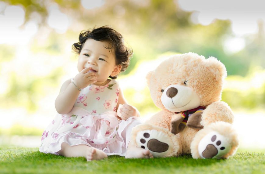 Smiling baby girl sits on grass with a plush teddy bear, enjoying a sunny day.