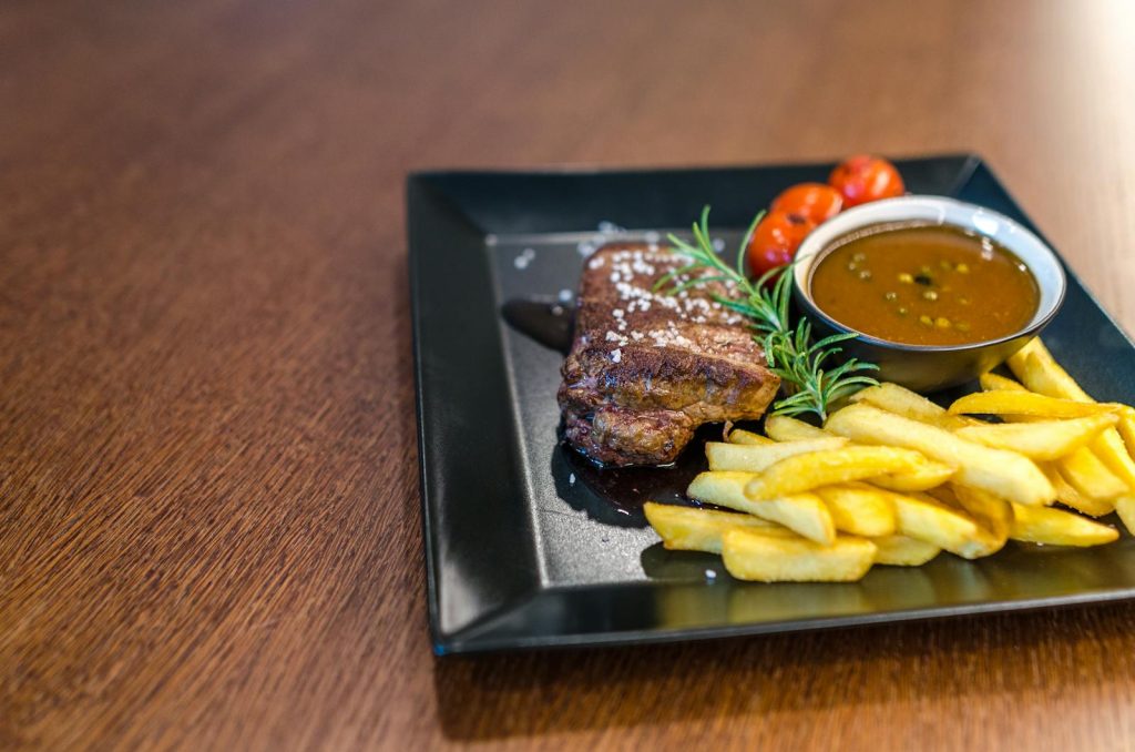 Close-up of a savory steak with fries, cherry tomatoes, and sauce on a black plate.