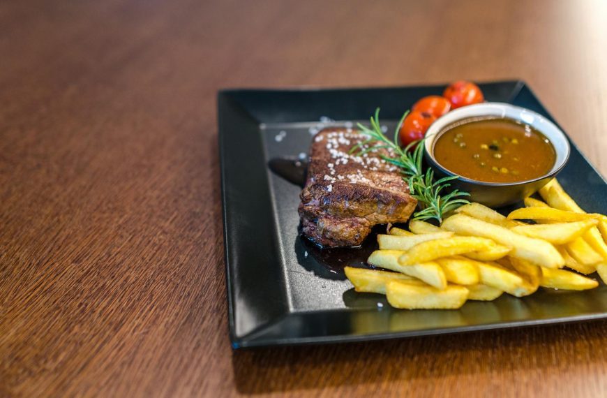 Close-up of a savory steak with fries, cherry tomatoes, and sauce on a black plate.