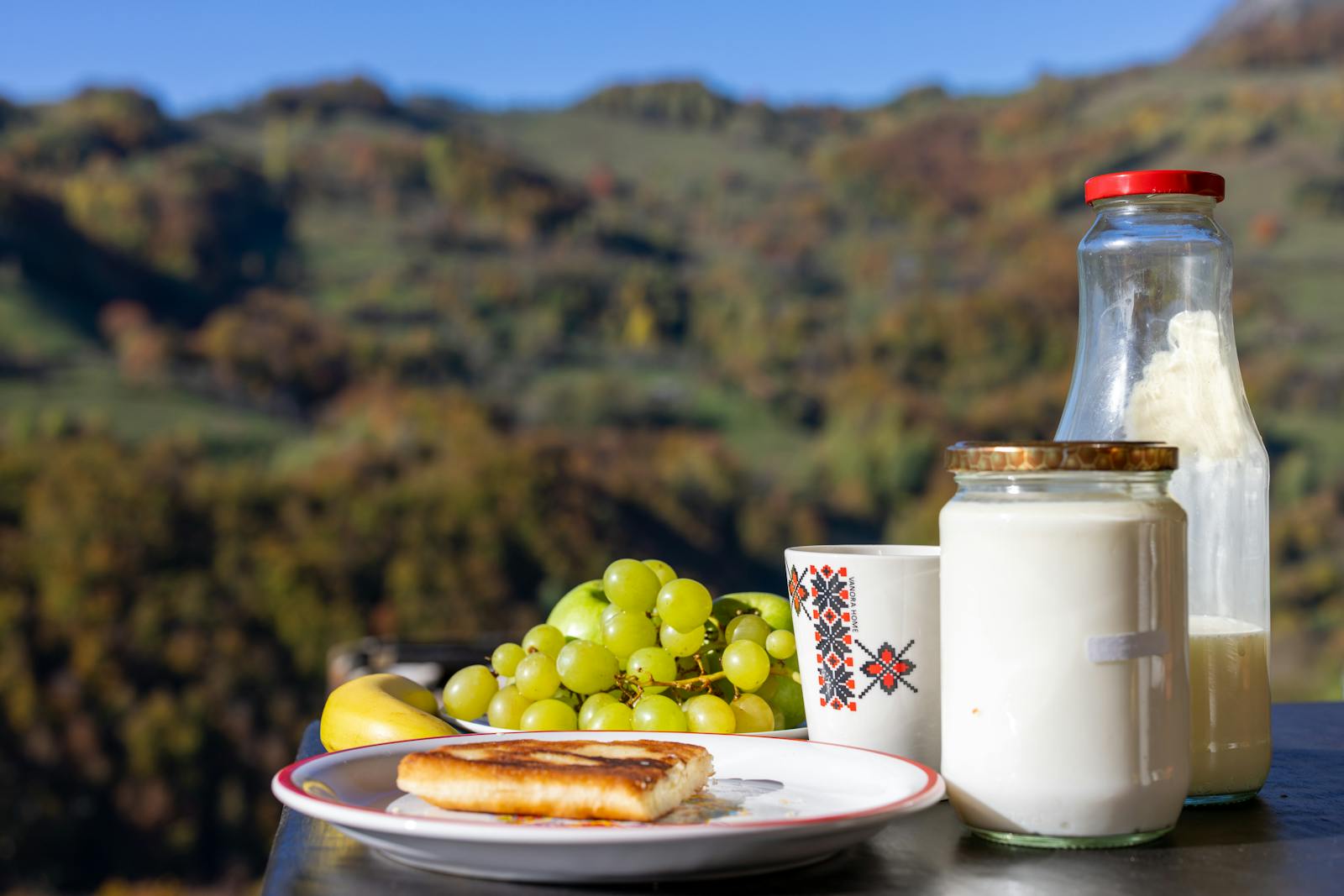 Scenic outdoor breakfast setup with milk, grapes, and pancake, set against a mountain backdrop.