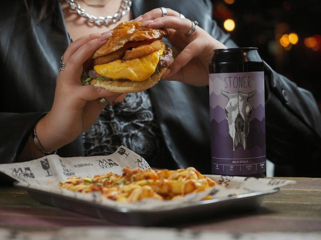 Close-up of a cheeseburger and beer can held by a person at a restaurant table.