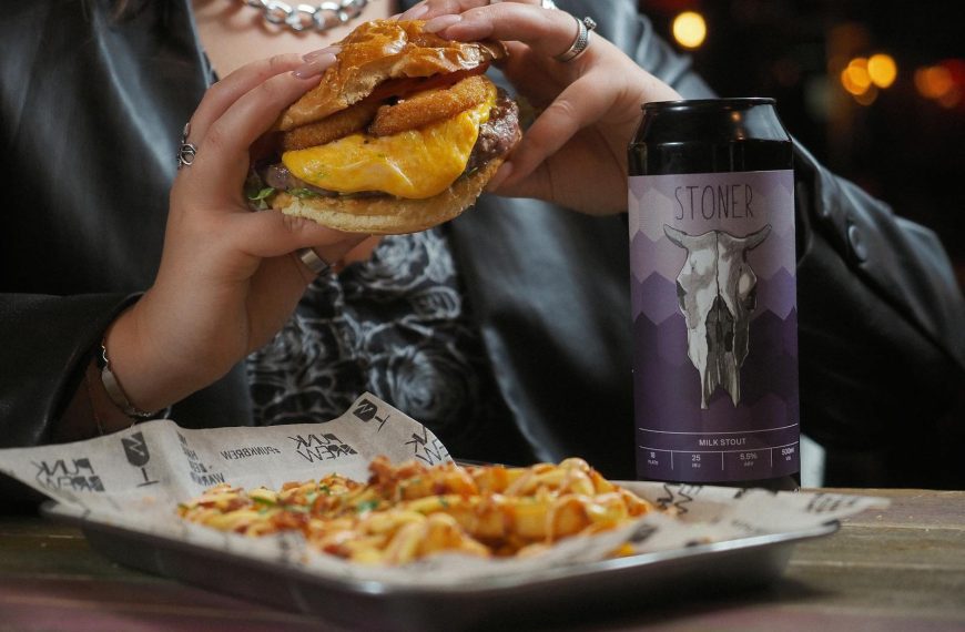 Close-up of a cheeseburger and beer can held by a person at a restaurant table.