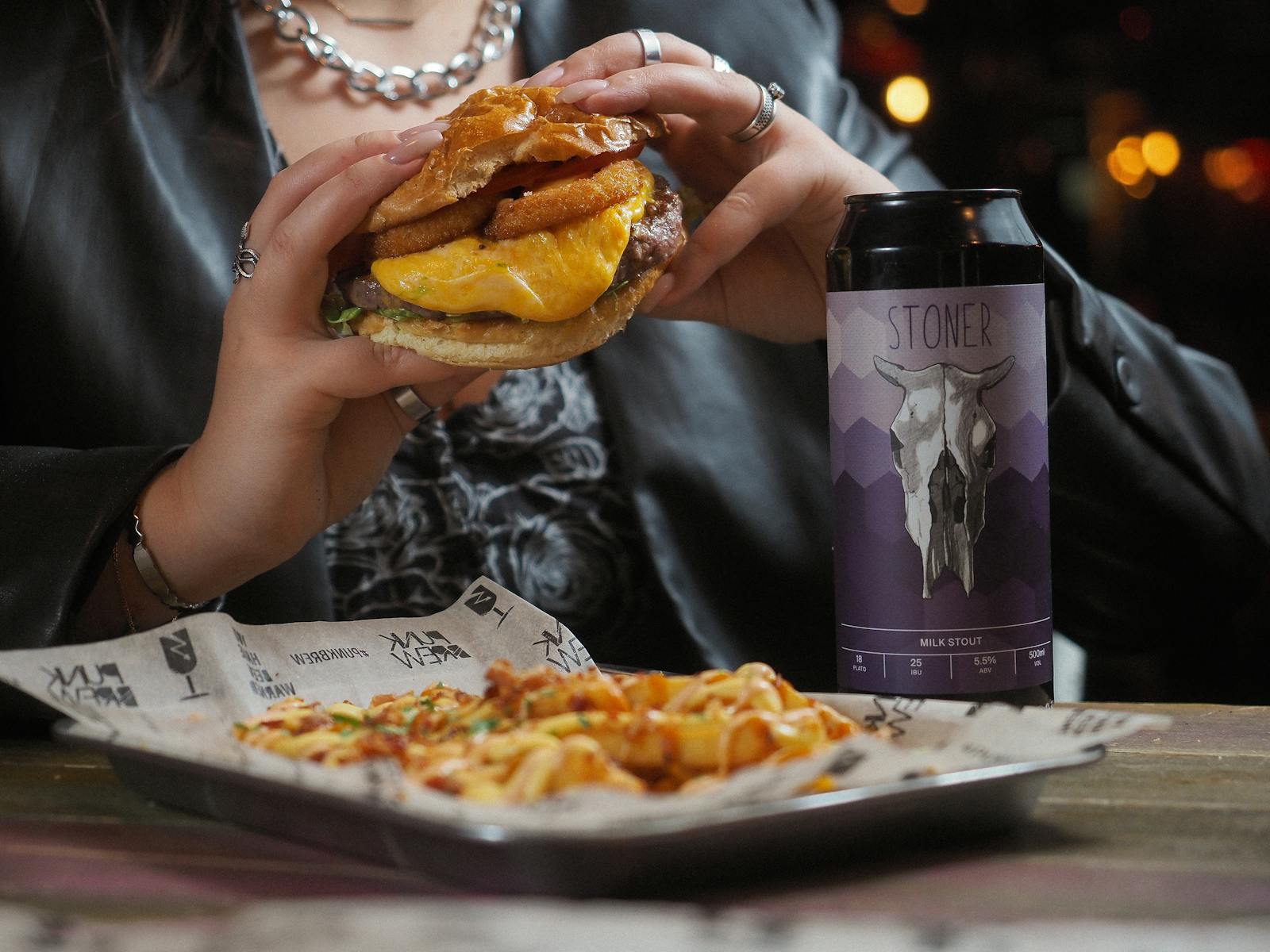 Close-up of a cheeseburger and beer can held by a person at a restaurant table.