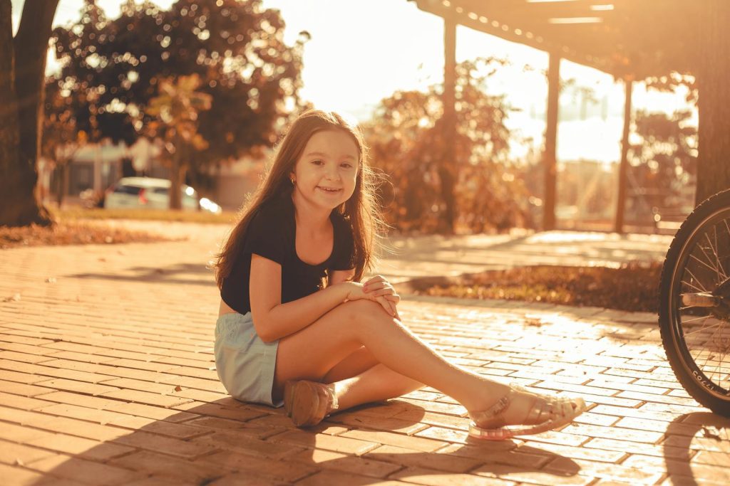 A young girl sits smiling on a sunlit sidewalk, exuding innocence and joy.
