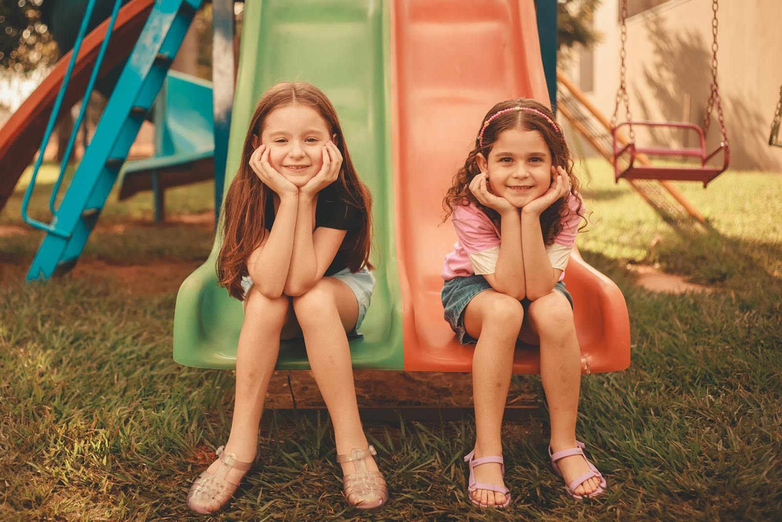 Two young girls sitting on a colorful playground slide, enjoying a sunny day outdoors.