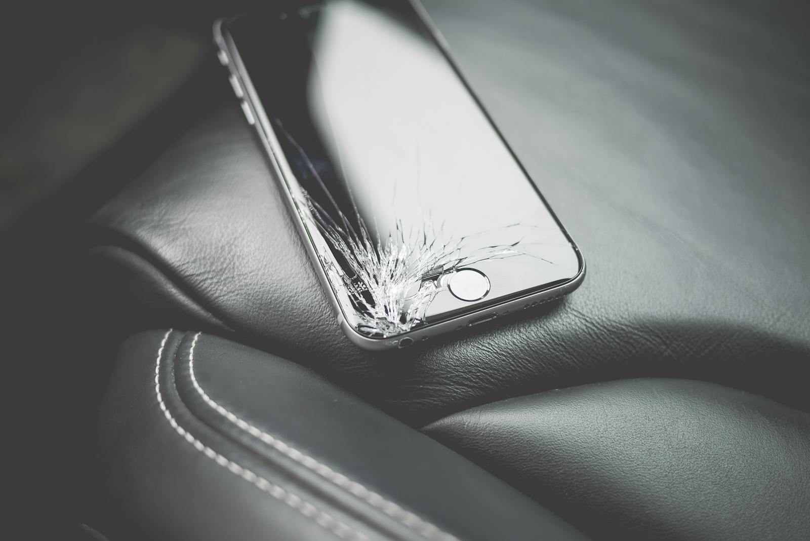 Close-up of a broken smartphone with a cracked screen on a leather car seat.