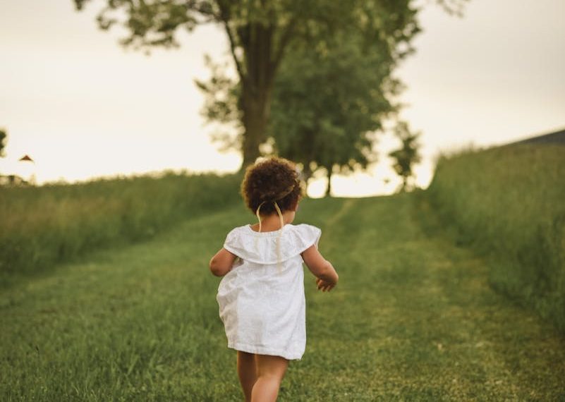 A young child runs freely down a grassy path surrounded by lush trees in a park during summer.