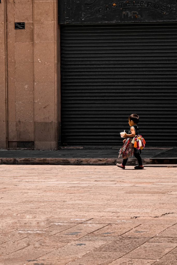 A child carrying a toy walks along an urban street in San Luis Potosí, Mexico, against a closed storefront.