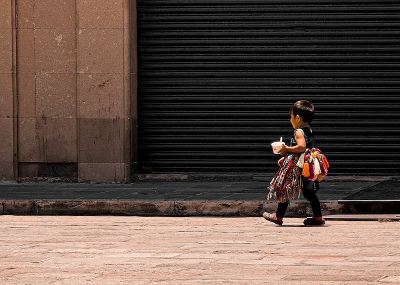 A child carrying a toy walks along an urban street in San Luis Potosí, Mexico, against a closed storefront.