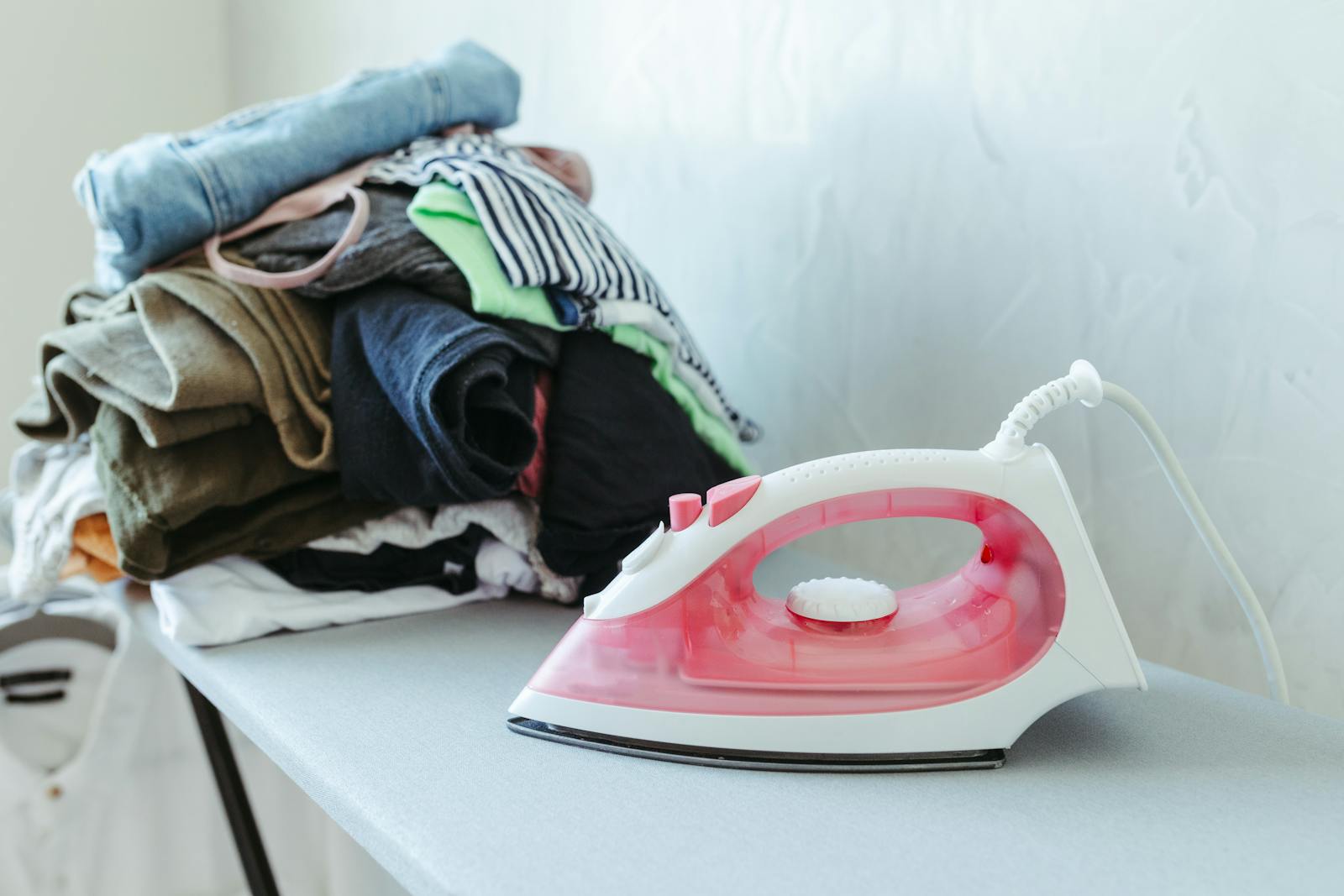 A red iron placed beside a stack of colorful clothes on an ironing board indoors.
