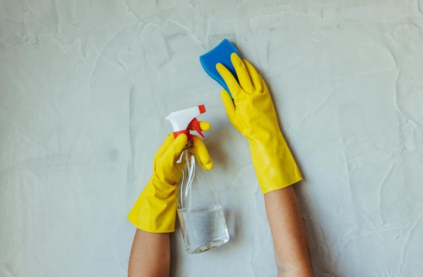 Close-up of hands in yellow gloves cleaning a wall with a spray bottle and sponge.