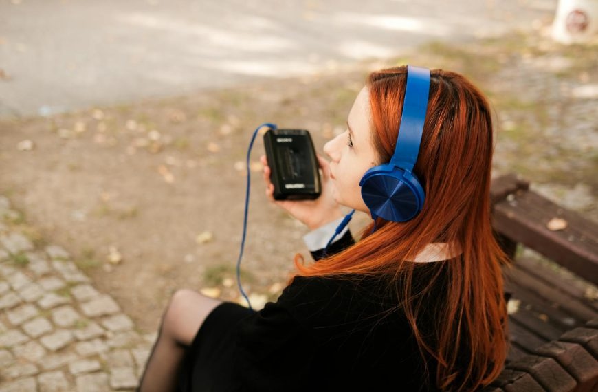 A redheaded teenager listens to music on headphones while sitting on a bench outside.