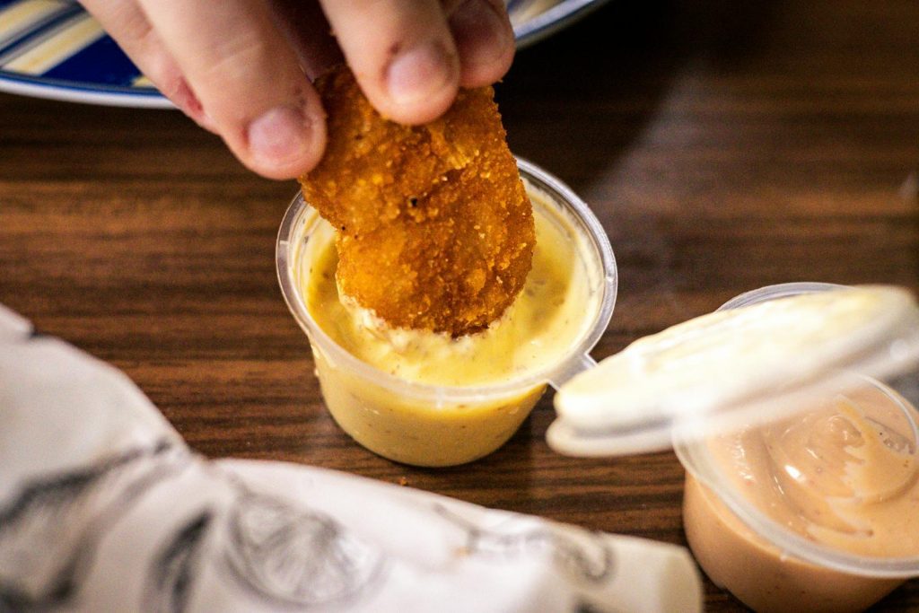 A crispy nugget being dipped into a cup of creamy yellow sauce, close-up view.