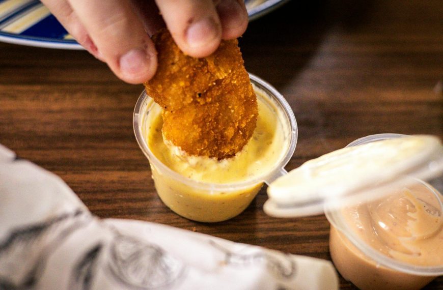 A crispy nugget being dipped into a cup of creamy yellow sauce, close-up view.