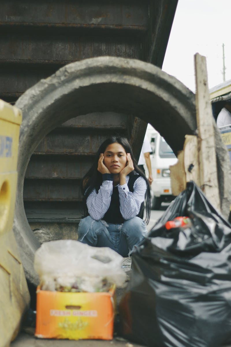 A woman sits amidst urban waste, emphasizing pollution and recycling issues.