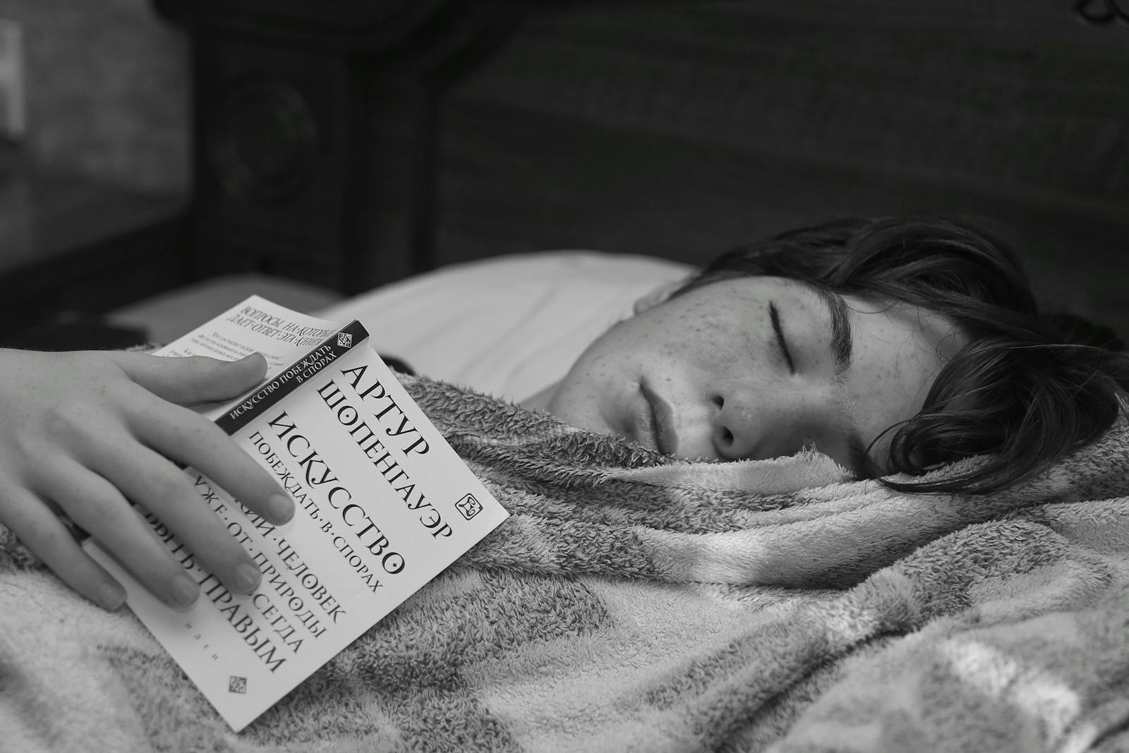 A teenage boy rests with a book in a cozy black and white setting.