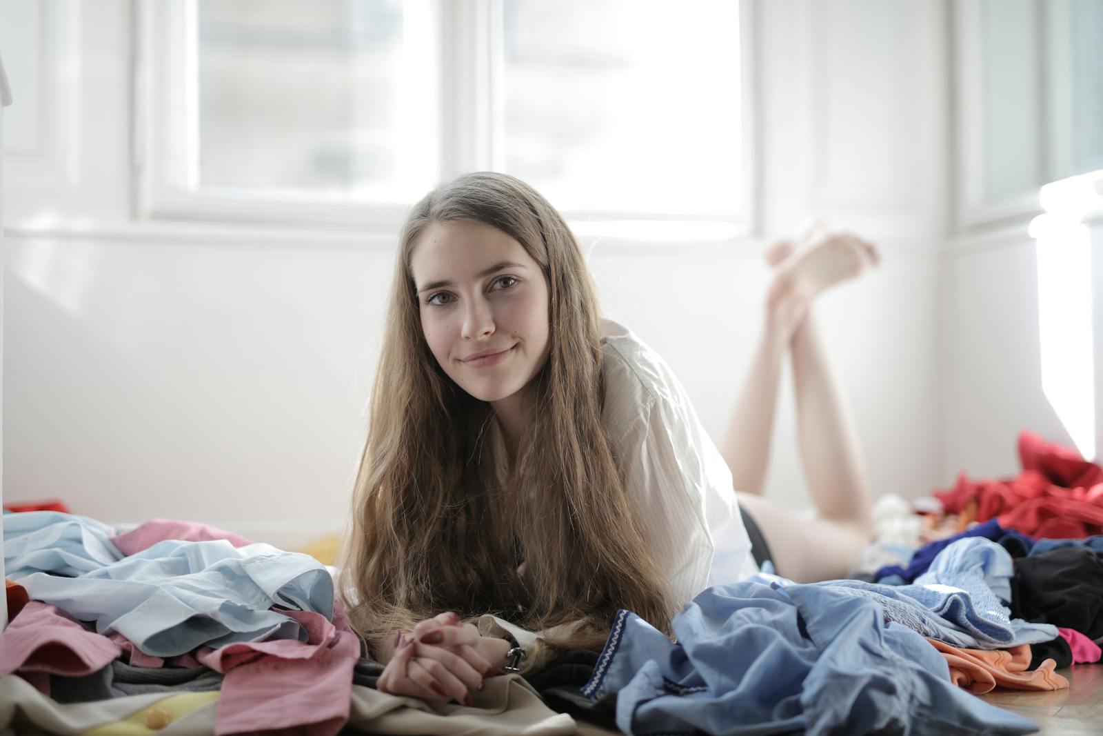 A young woman with a smile lying comfortably amidst colorful clothes in a sunny bedroom.