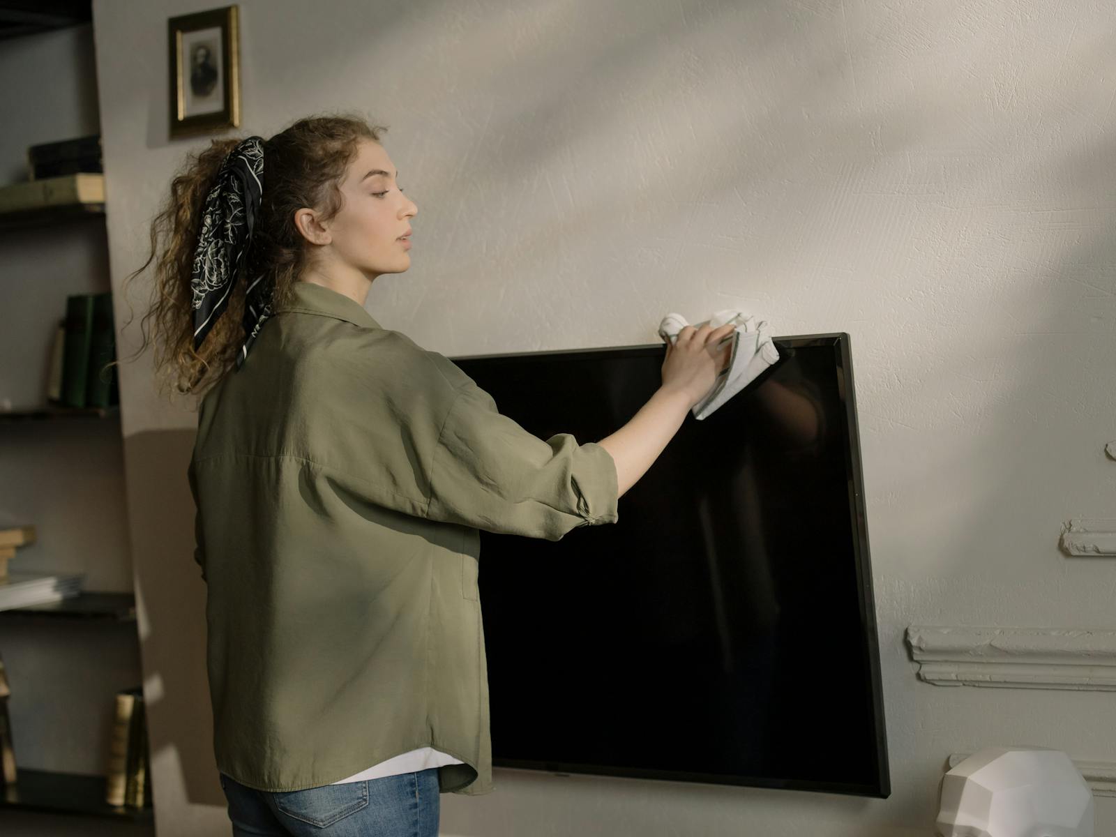 Woman with curly hair cleans a TV screen in a cozy home, creating a clean and tidy living space.