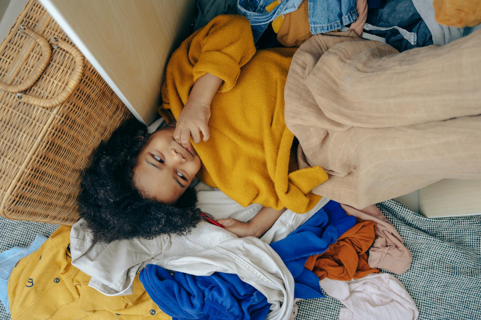 Adorable child lying contentedly in a pile of colorful cozy clothes indoors.