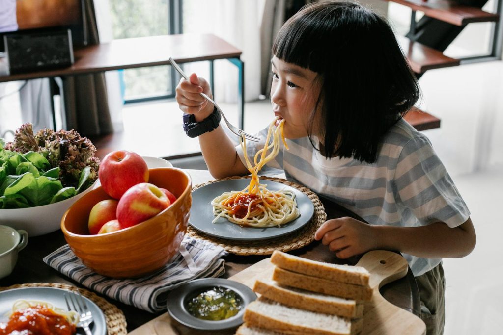 A child happily eats homemade spaghetti at a table with fresh apples and bread, creating a cozy indoor dining scene.