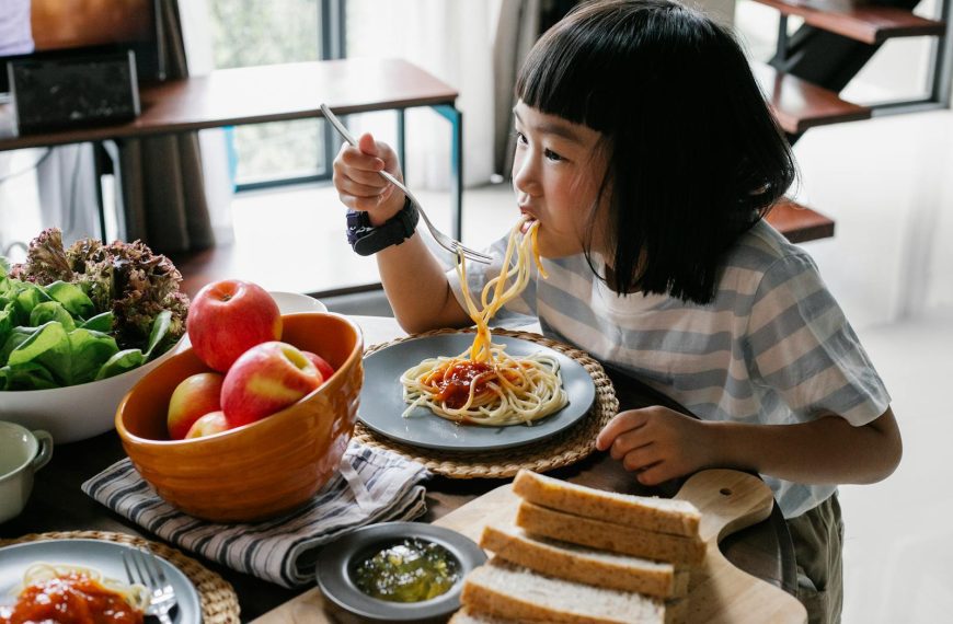 A child happily eats homemade spaghetti at a table with fresh apples and bread, creating a cozy indoor dining scene.