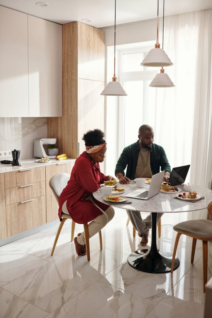 A couple enjoys breakfast while using laptops in a modern kitchen setting during the morning.