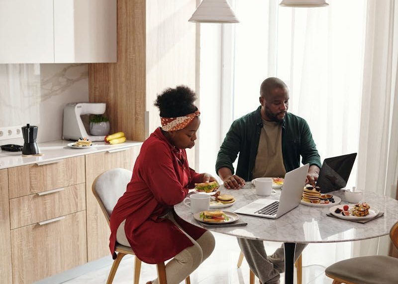 A couple enjoys breakfast while using laptops in a modern kitchen setting during the morning.