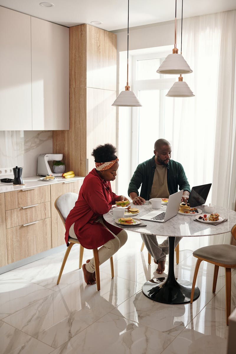 A couple enjoys breakfast while using laptops in a modern kitchen setting during the morning.