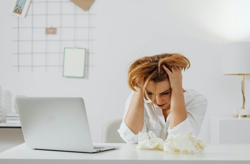 Frustrated woman in white shirt at desk with laptop, feeling overwhelmed.