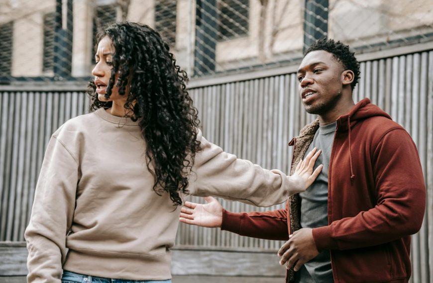 A couple in casual clothing appears to be in a disagreement outdoors with grey fencing background.