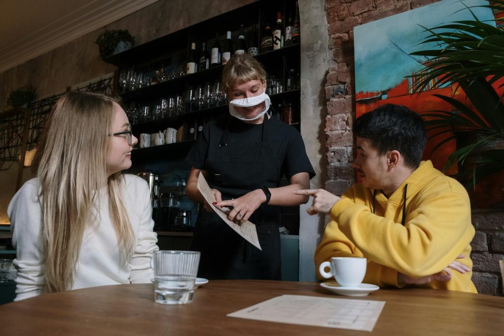 A masked waitress serves two patrons in a cozy restaurant setting.