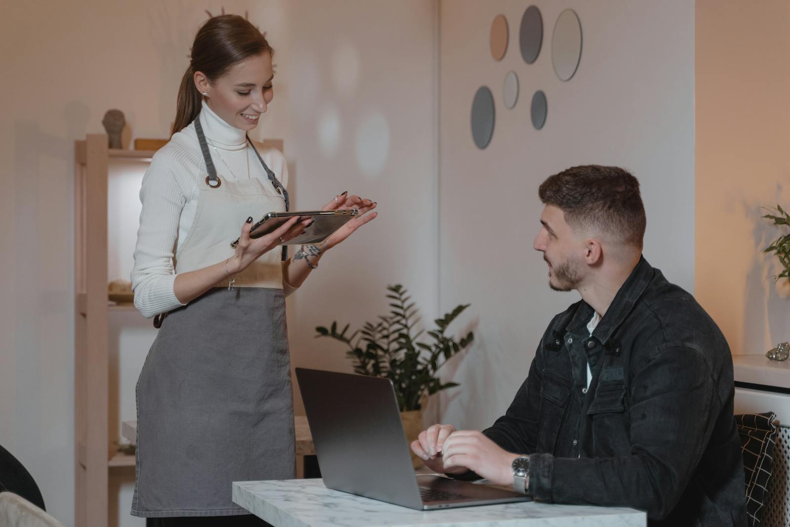 A smiling waitress takes an order from a customer on a tablet in a cozy café setting.