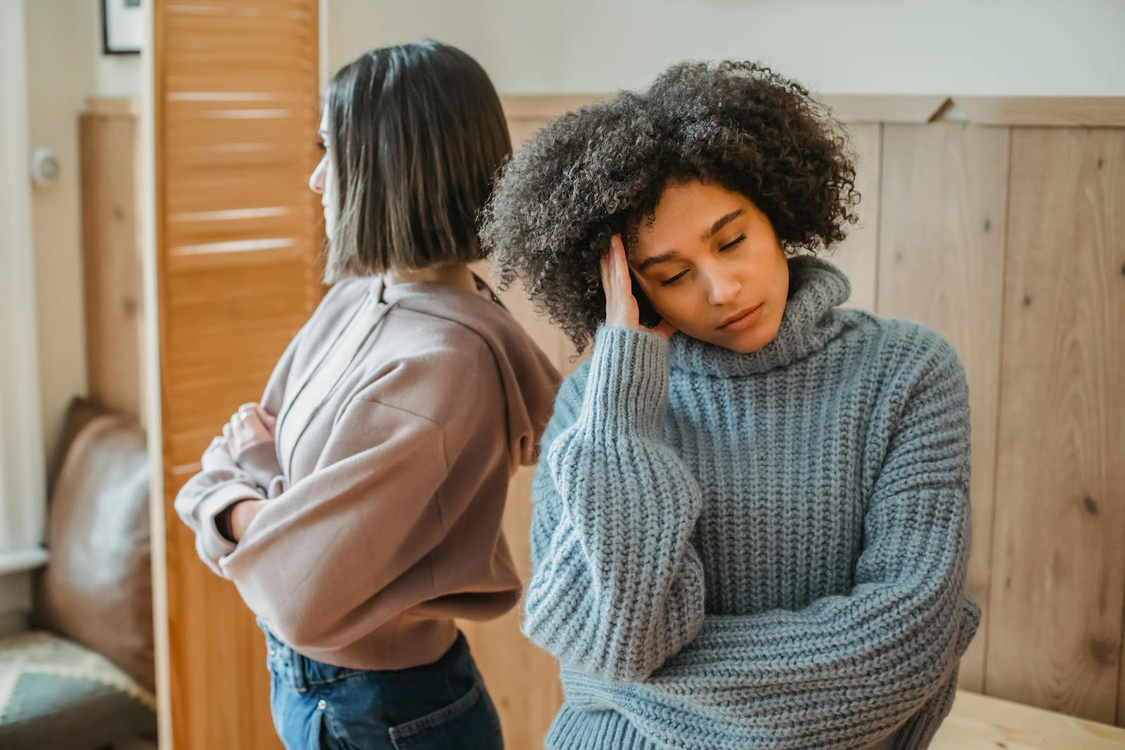 Upset African American woman with curly hair and offended female standing in light room with wooden wall while having conflict