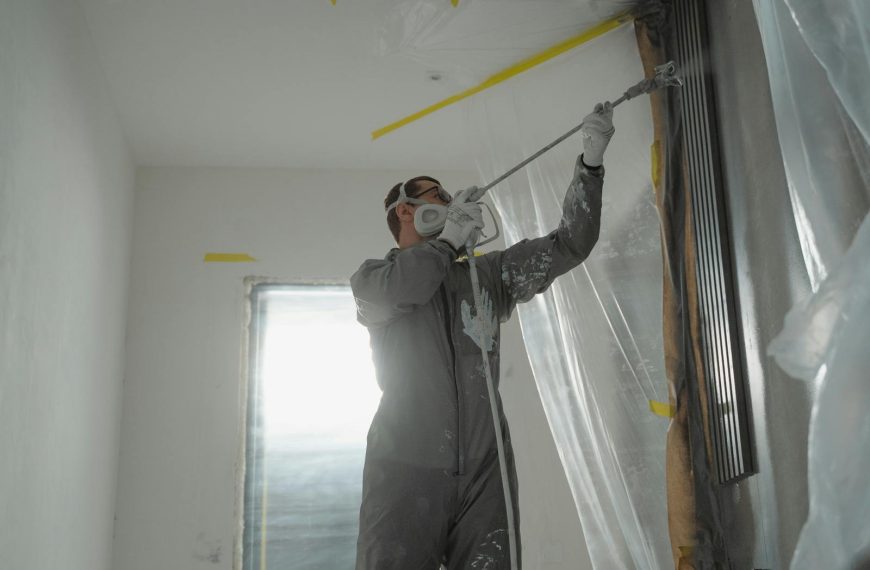 A professional painter wearing protective gear spray paints a wall indoors, working on a home renovation project.