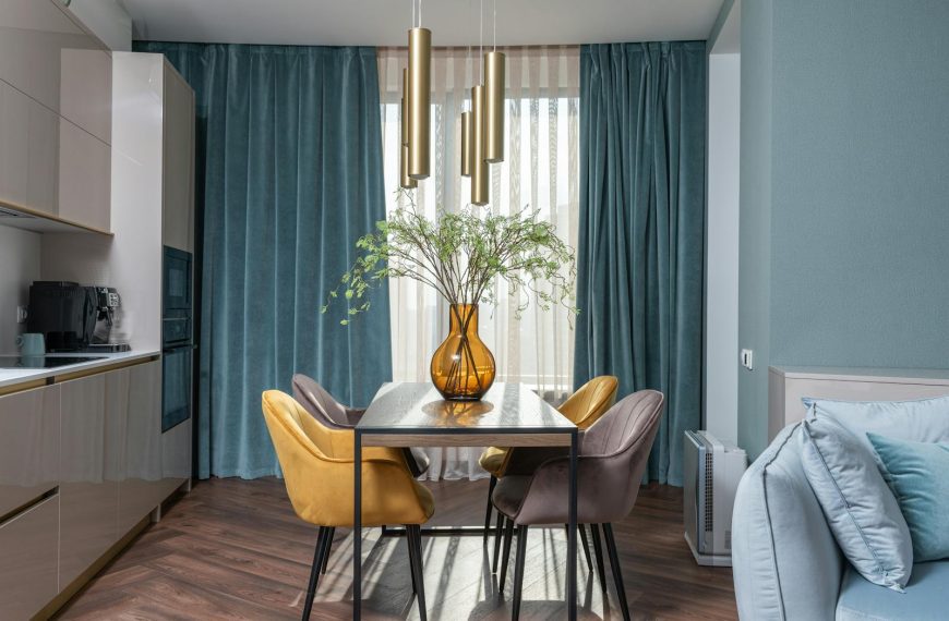 Glass vase with flowers on table with chairs placed near kitchen cabinets and soft couch in modern apartment with window