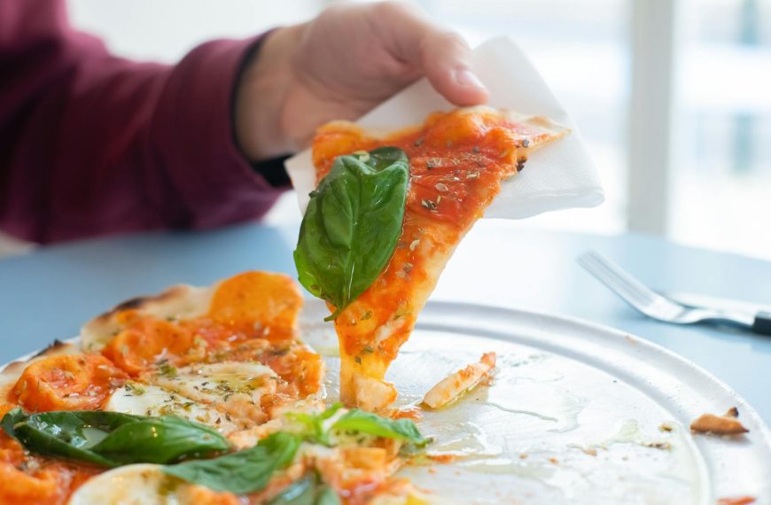 Close-up of a hand holding a slice of delicious Margherita pizza with basil topping on a sunny day.