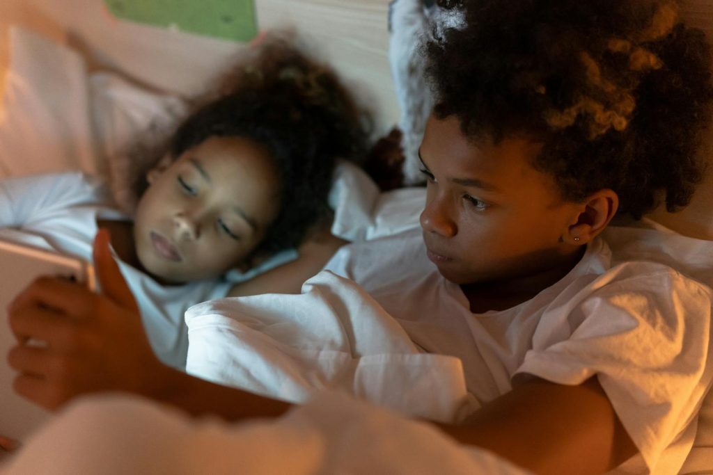 Two children in pajamas lying in bed, engaging with a tablet screen under cozy lighting.