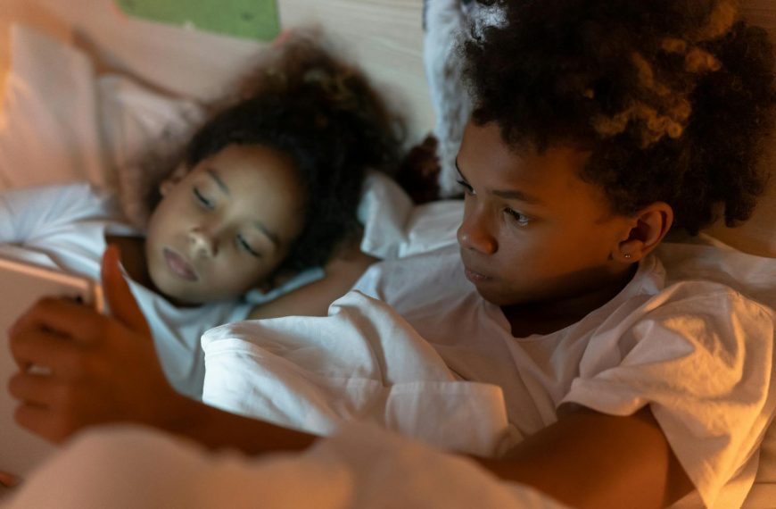 Two children in pajamas lying in bed, engaging with a tablet screen under cozy lighting.