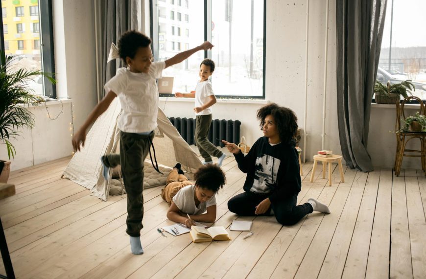 Full body of African American mother sitting on wooden floor near daughter writing in copybook in light room with playing boys