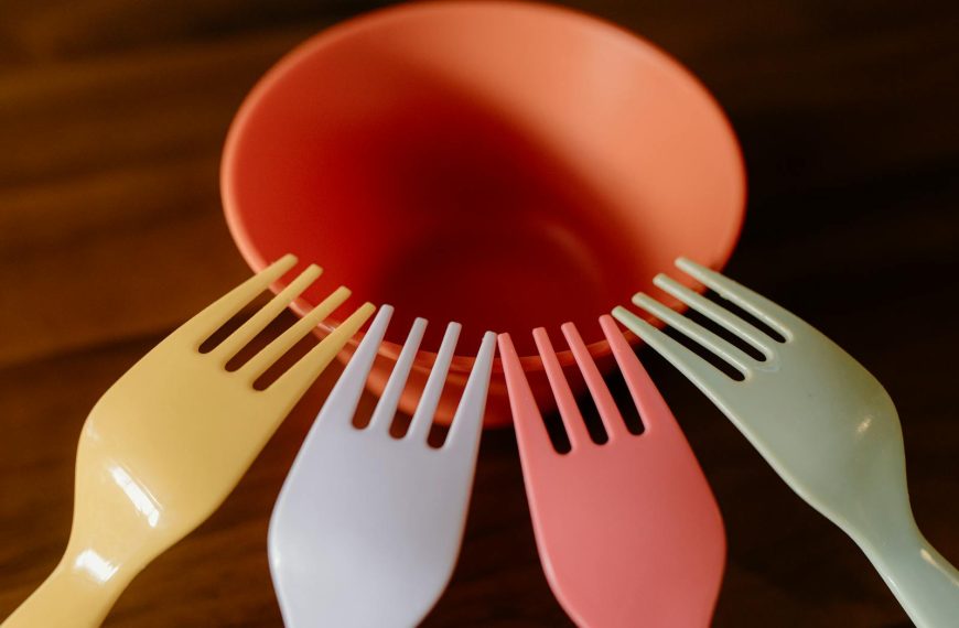 A vibrant close-up of colorful plastic forks surrounding an orange bowl on a wooden table.