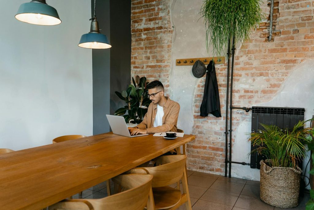 Man working on laptop in a stylish indoor setting with brick wall and plants.