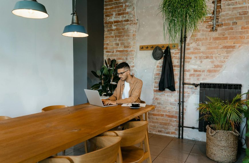 Man working on laptop in a stylish indoor setting with brick wall and plants.