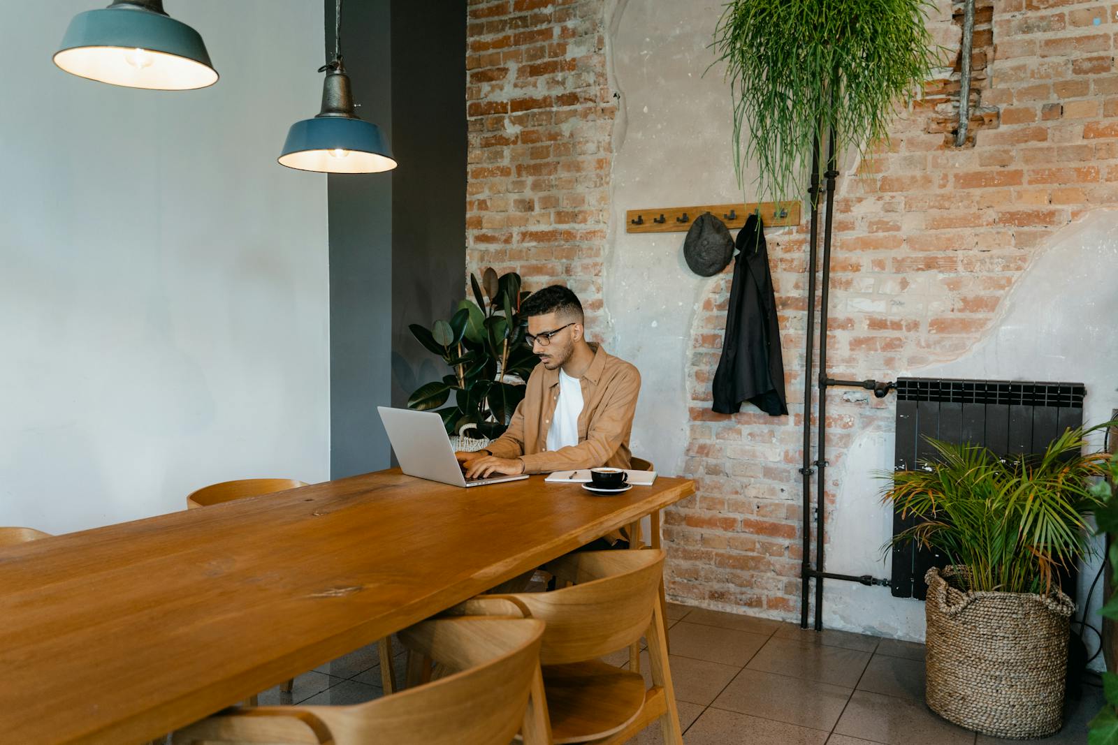Man working on laptop in a stylish indoor setting with brick wall and plants.